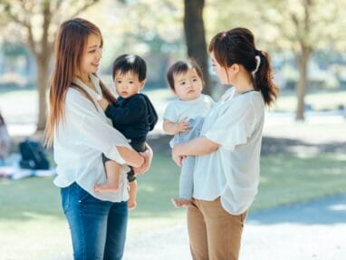 4329539_s | 保育士求人|大阪・兵庫・関西の保育士・幼稚園教諭の就職転職支援|保育のせかい Two women in white tops hold young children and chat in a sunny park, smiling at each other.